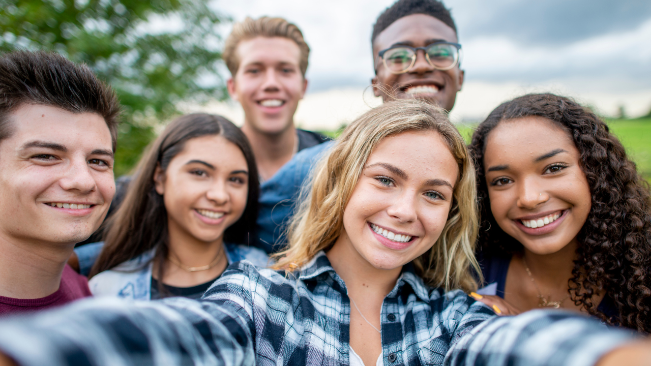 Six smiling teenagers taking a selfie together outdoors, with trees, grass, and a cloudy sky in the background.